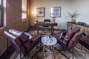 Office space with two burgundy leather chairs separated by marble side table, desk workspace, and brick accent wall