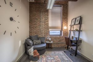 Waiting area with gray curved sofa, minimalist wall clock, exposed brick, window with blinds, and shelving unit