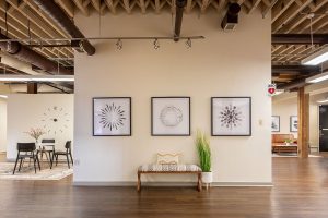 Open waiting area with geometric wall art, wooden bench seating, plants, and industrial ceiling