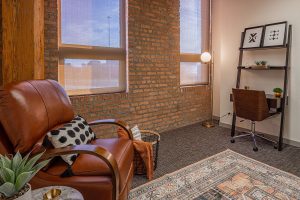 Therapy office with cognac leather armchair, brick accent wall, large windows with city views, and desk with shelving