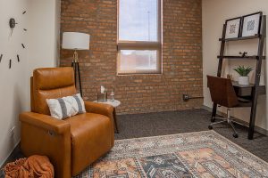Individual therapy room with tan leather reclining chair, brick wall, window with natural light, and desk with shelving