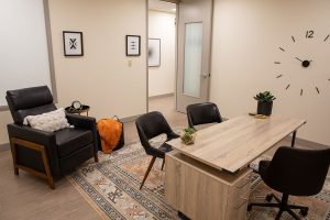 Clinical consultation room with black recliners, wooden desk, chairs, and wall clock at Relief Mental Health