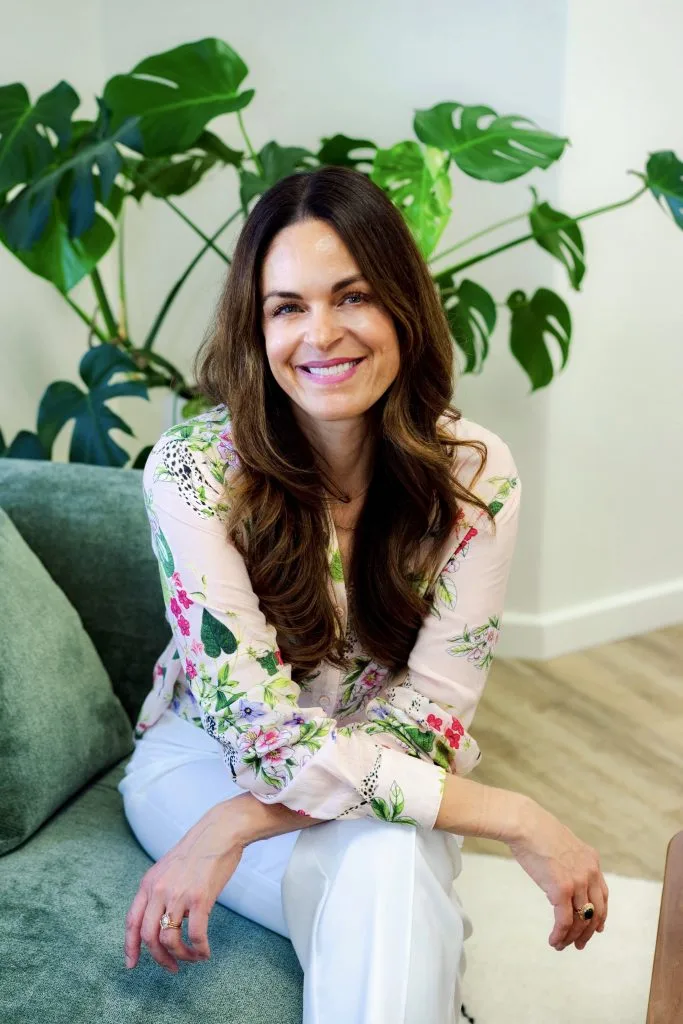 Rosina, staff member, sitting on green chair wearing floral blouse with Monstera plant behind her