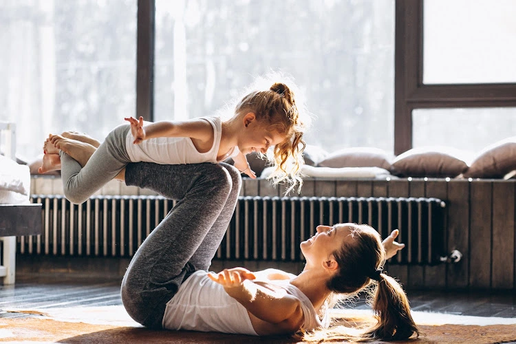 Woman and child doing partner yoga pose on floor near large windows in bright studio