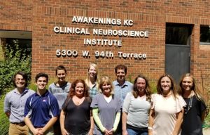 Team of ten staff members standing in front of red brick building with white signage reading 'AWAKENINGS KC CLINICAL NEUROSCIENCE INSTITUTE 5300 W. 94th Terrace'