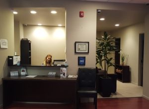 Medical clinic reception desk with black front counter, computer, and plant in warmly lit interior space