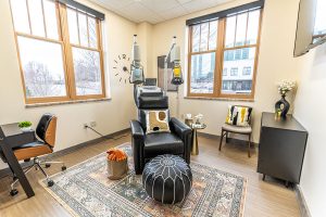 TMS therapy treatment room with black reclining chair, overhead TMS coils with yellow and gray casings, desk, and windows overlooking city