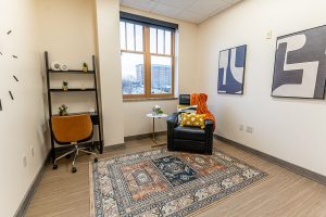 Private therapy office with black leather chair, wooden shelf unit, small table, geometric wall art, and city view from window