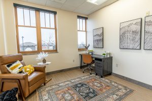 TMS therapy office lounge with brown leather chair, windows overlooking parking lot, desk with artwork, and patterned rug
