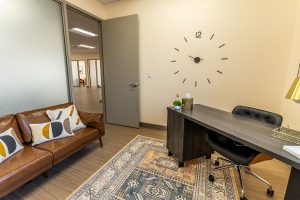 TMS therapy consultation room with brown leather sofa, wall clock, desk with chair, and frosted glass door