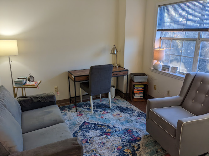 Therapy office with two gray armchairs, desk with chair, lamp, and large windows overlooking trees