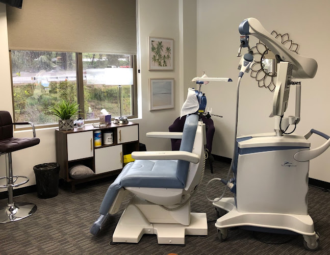 TMS treatment room with patient chair, overhead magnetic coil device, and window overlooking landscaped grounds