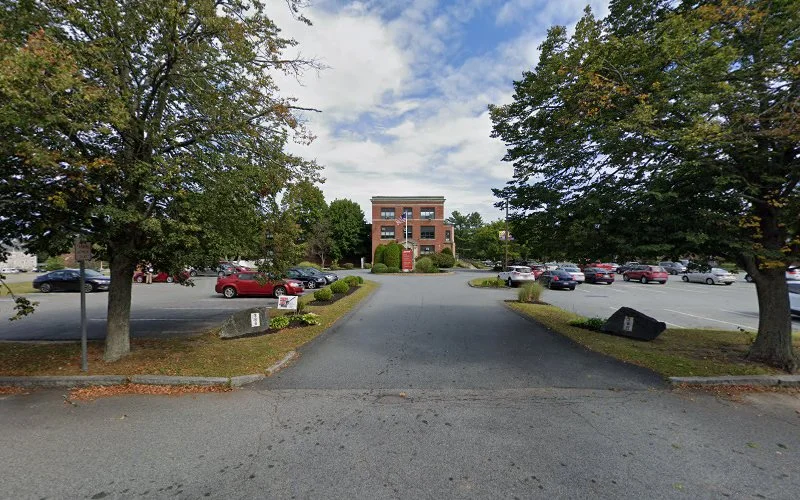 Red brick institutional building set back from parking lot lined with mature trees
