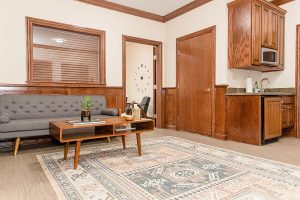 Patient lounge with gray upholstered sofa, wooden coffee table, kitchenette, and analog wall clock