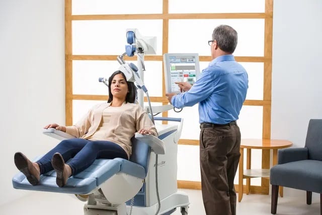 Clinician operating TMS device on seated patient in treatment room with wood-framed windows
