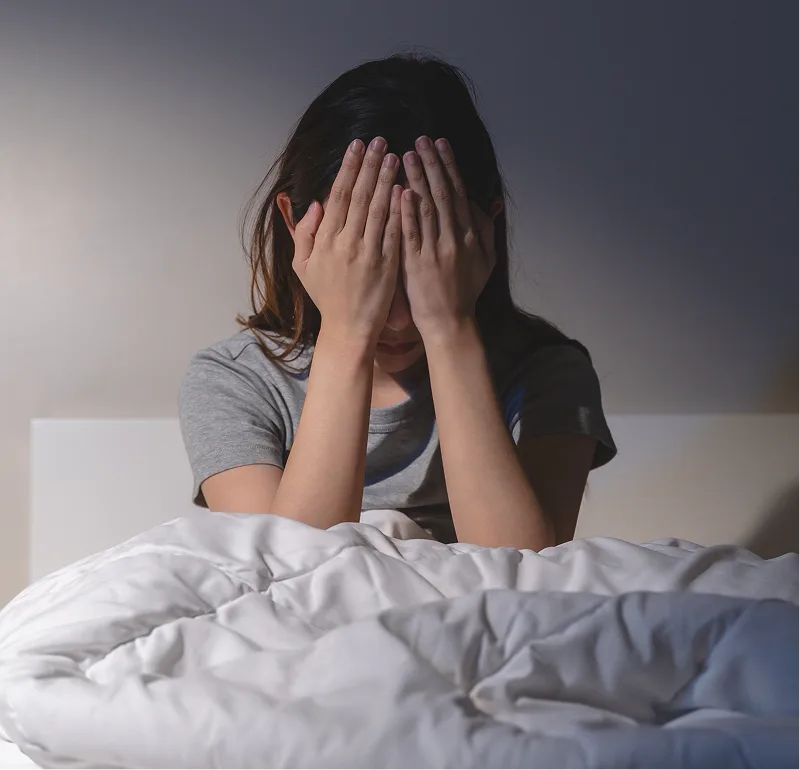 Woman sitting on bed covering her face with hands, experiencing sleep distress