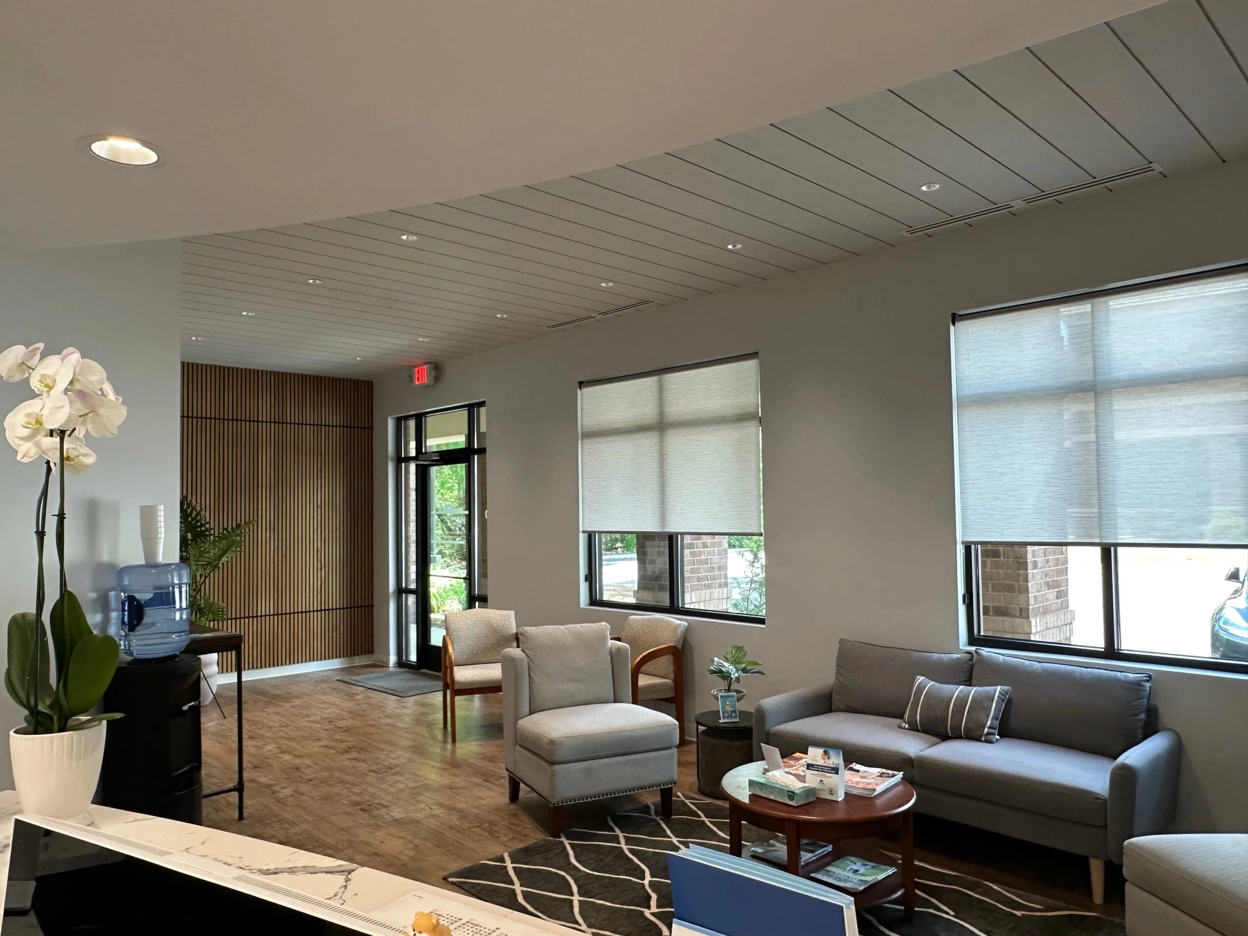 Modern waiting room with gray seating, wooden divider, orchid plant, windows with blinds, and recessed lighting