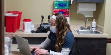 Medical staff member in mask typing on laptop at clinic desk with supplies and sink