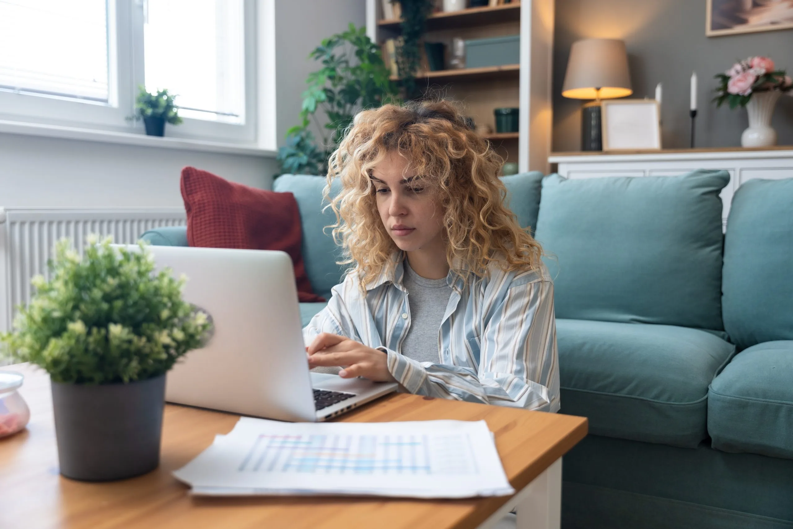 Woman with curly blonde hair working on laptop at home desk with papers and plant, teal couch and shelving behind her
