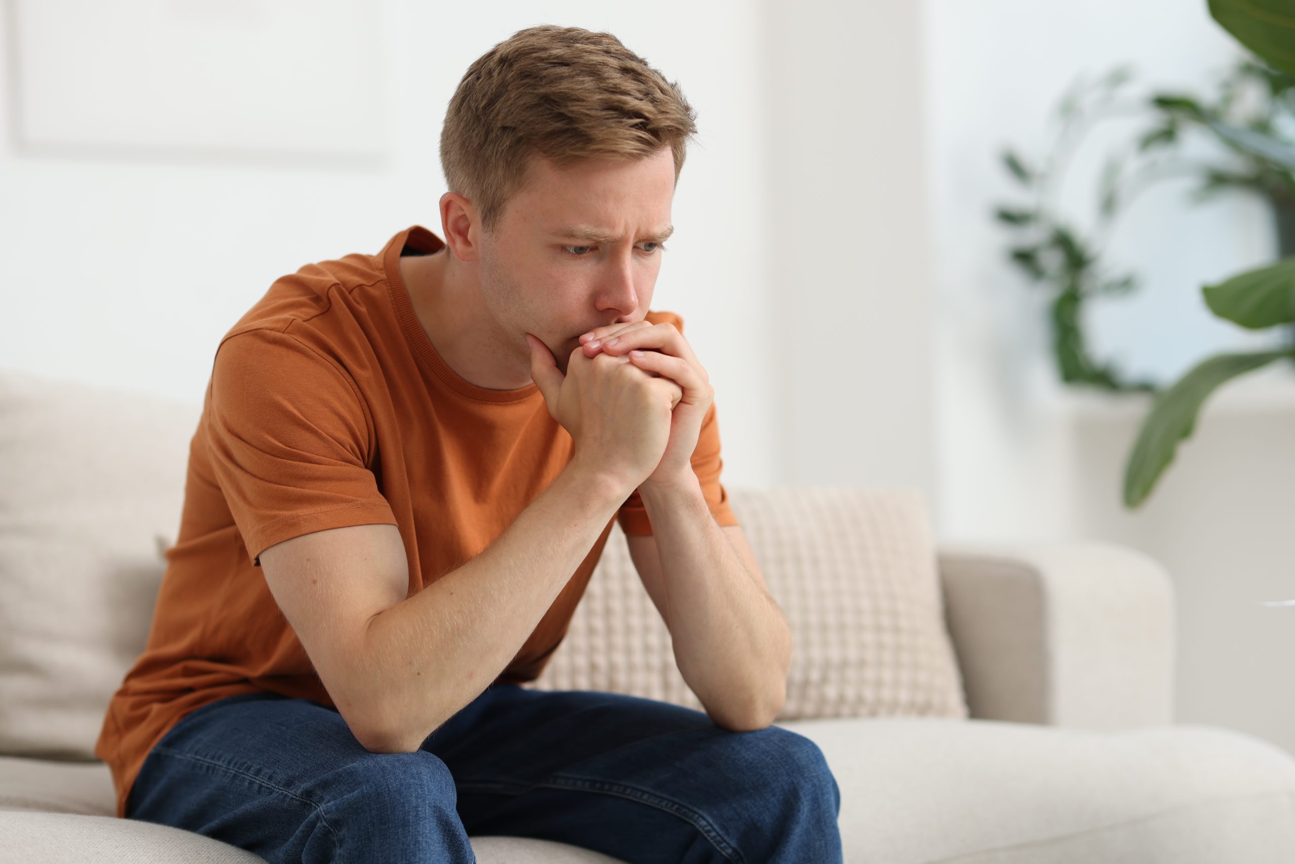 Man in orange shirt sitting on couch with hands clasped near face, looking troubled