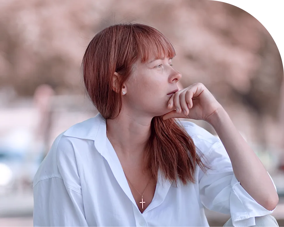 Woman with reddish-brown hair in white shirt, chin resting on hand, gazing thoughtfully to the side