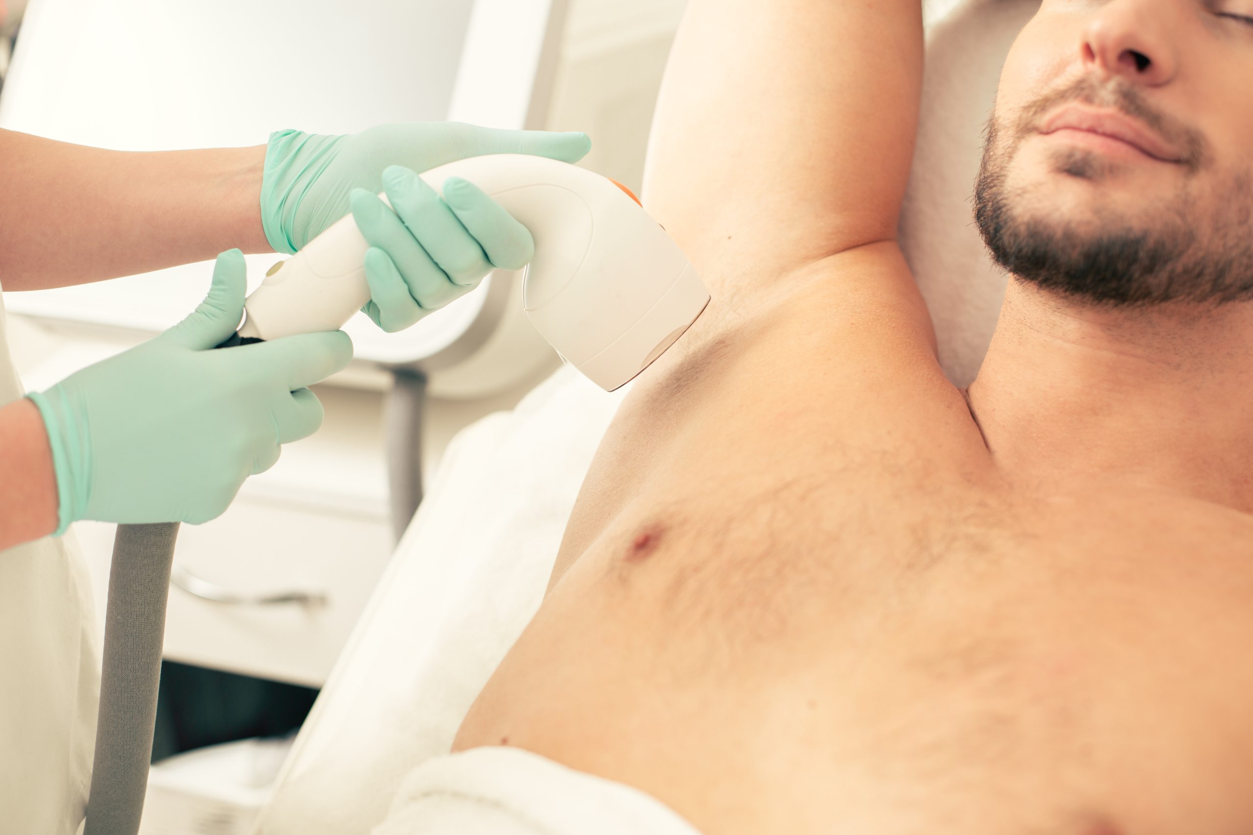 Provider in green gloves using laser device to remove hair from man's underarm