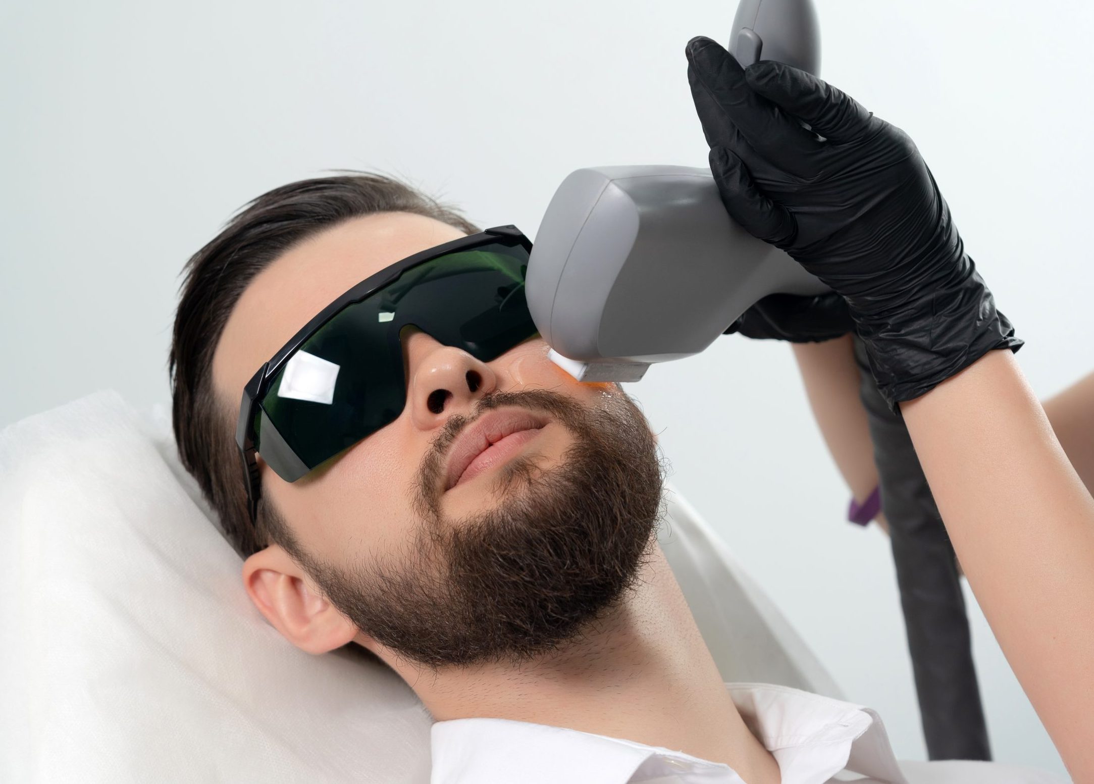 Bearded man receiving laser facial treatment from cosmetologist wearing protective eyewear and black gloves