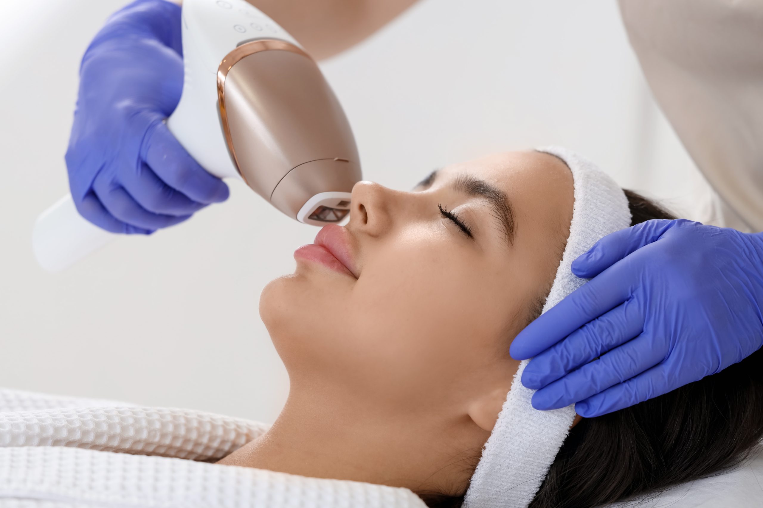 Aesthetician using handheld laser device to remove facial hair from woman's upper lip in salon treatment