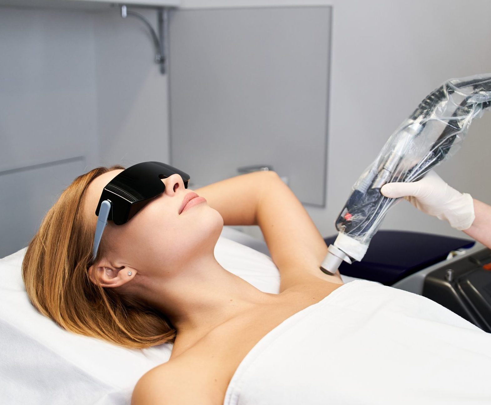 Woman wearing protective eyewear relaxes while technician applies laser hair removal device to underarm