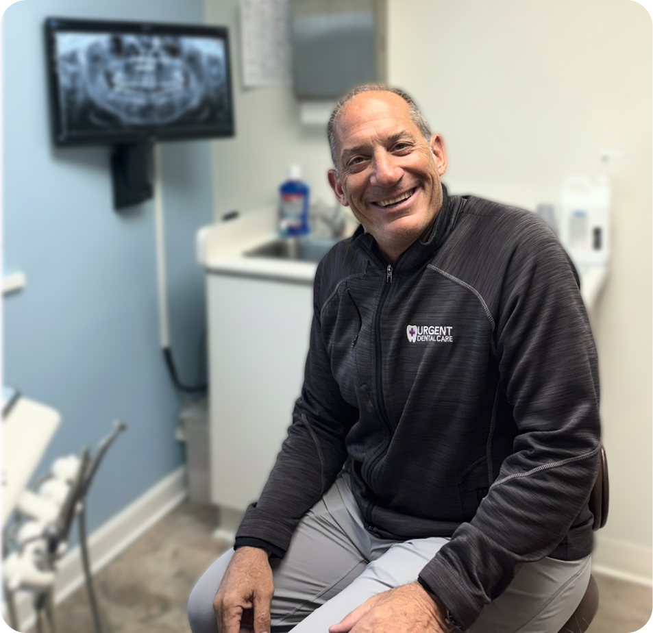 Dentist in black Urgent Dental Care jacket smiling in treatment room with x-ray displayed on wall monitor