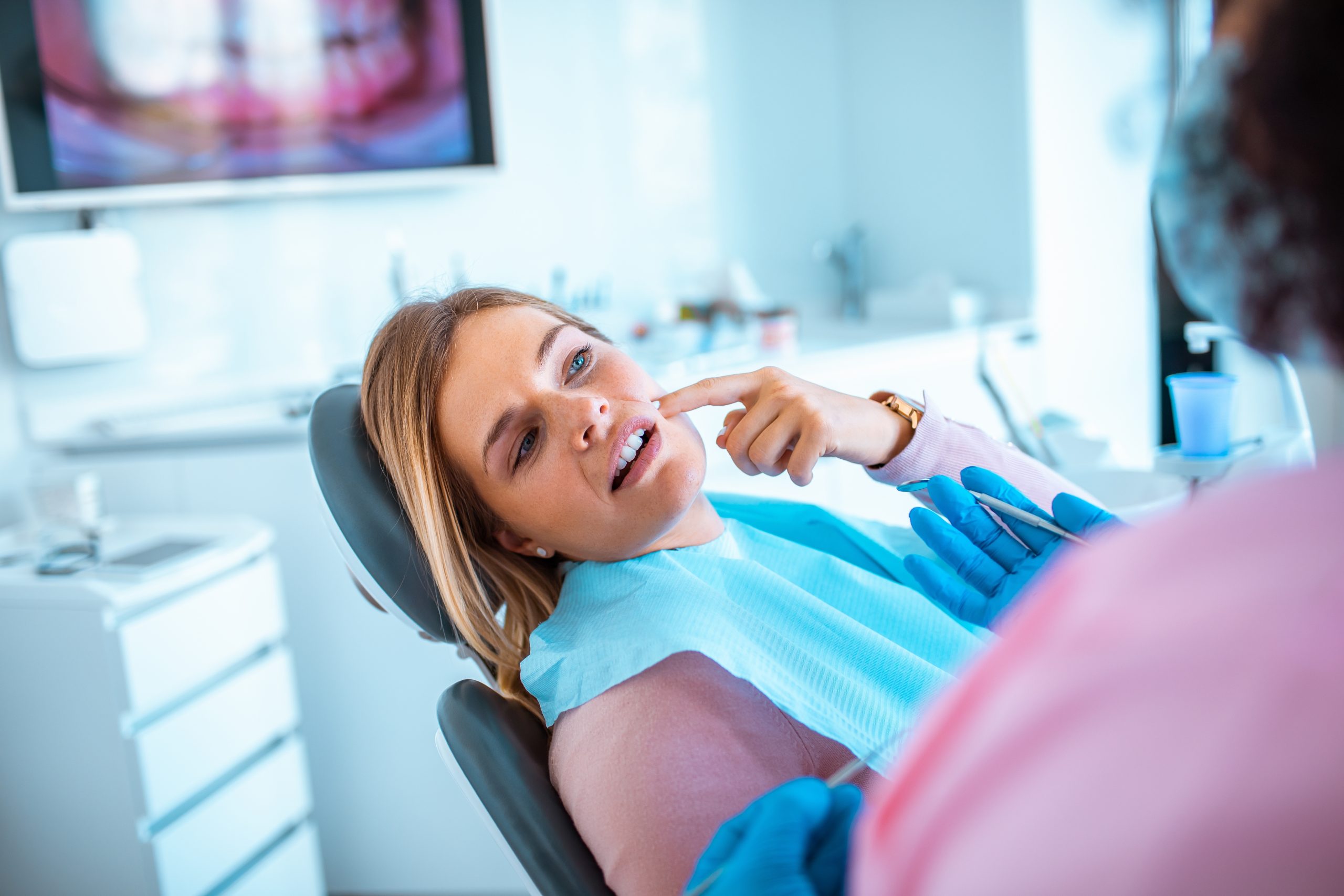 Dentist in blue gloves examining patient's teeth in dental chair with monitor displaying dental imagery