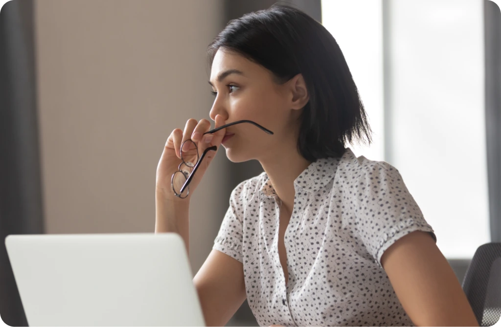 Woman holding her glasses and looking thoughtfully out a window while sitting at a laptop