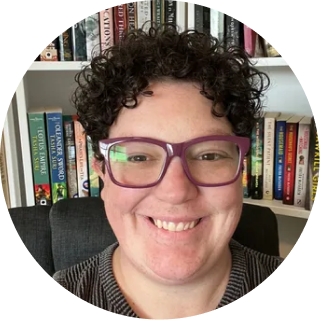Person wearing purple glasses and dark curly hair, smiling in front of a bookshelf