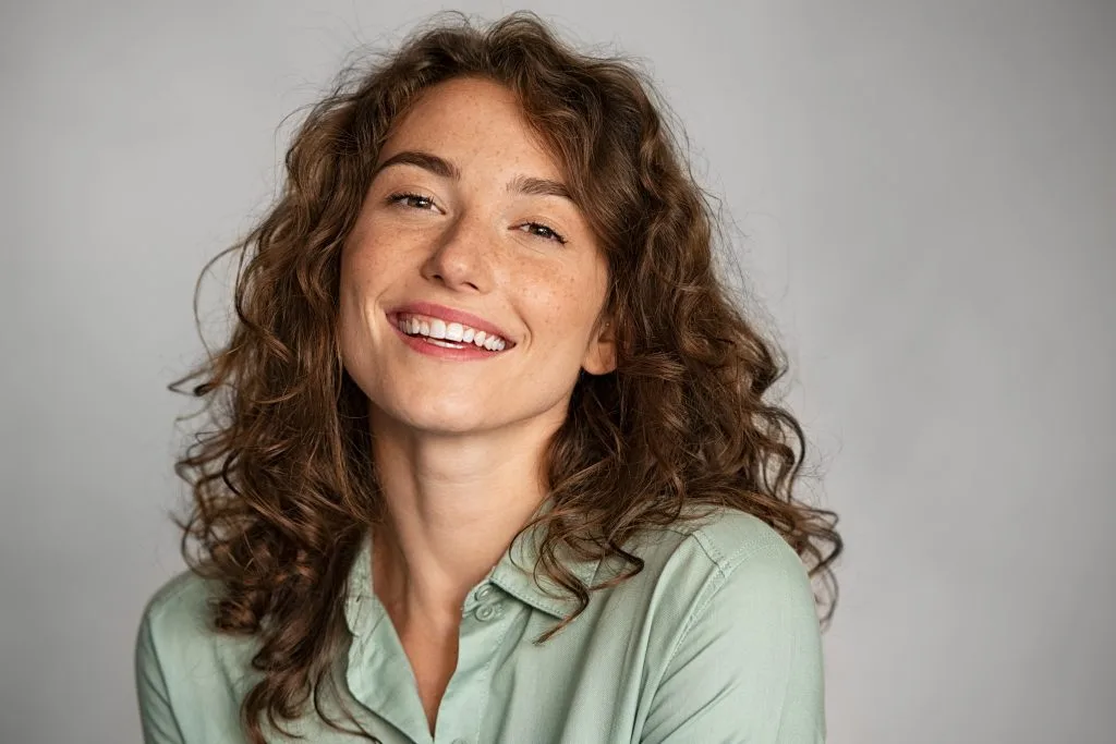 Woman with curly brown hair smiling against neutral gray background