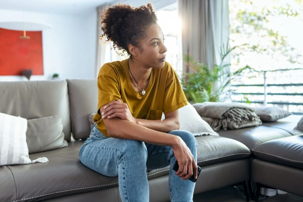 Woman in yellow shirt sitting on couch looking thoughtfully to the side in bright living room