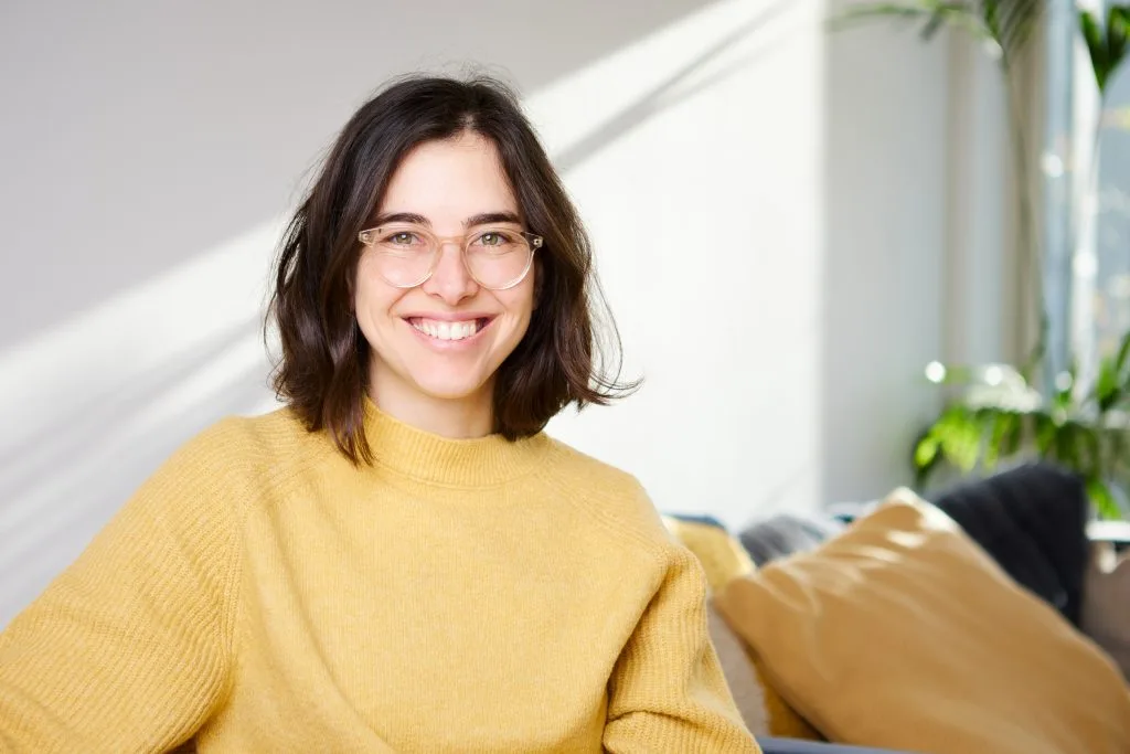 Woman wearing yellow sweater and glasses smiling in bright sunlit room with green plants