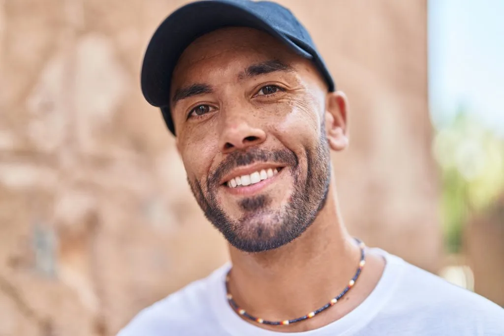 Man wearing blue cap and beaded necklace smiling outdoors against brick wall