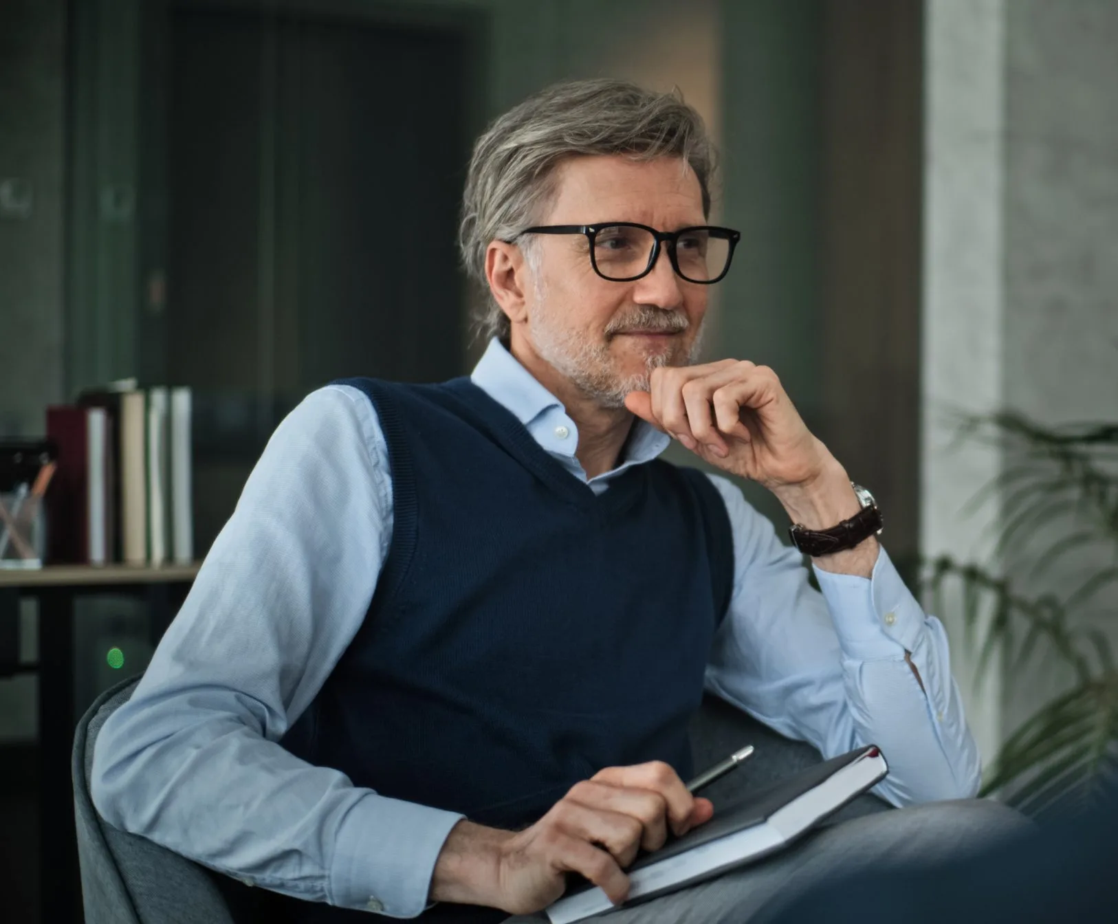 Therapist in glasses holding notepad, thoughtfully posed at desk with bookshelves behind