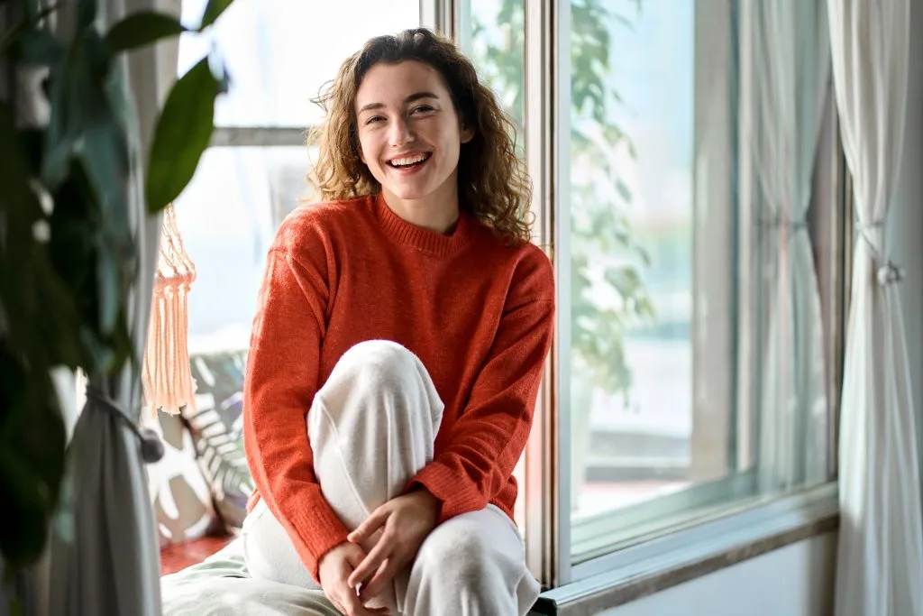 Woman in red sweater smiling by sunny window surrounded by potted plants