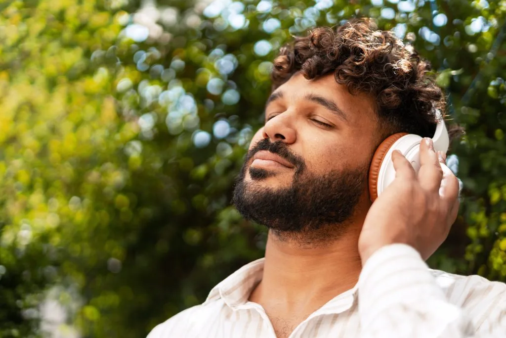 Man with headphones closed eyes listening to music outdoors among green foliage