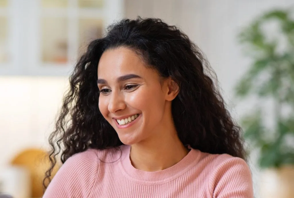 Woman with curly dark hair in pink sweater smiling in bright home setting