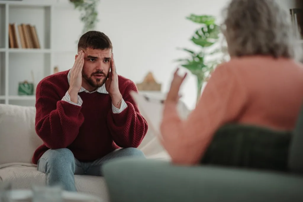 Man in maroon sweater holds head in distress during therapy session with counselor