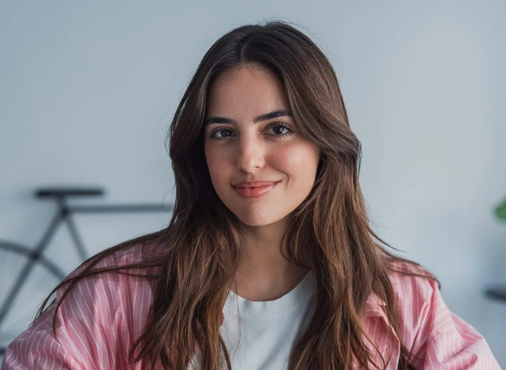 Woman with long brown hair in pink striped shirt smiling at camera indoors