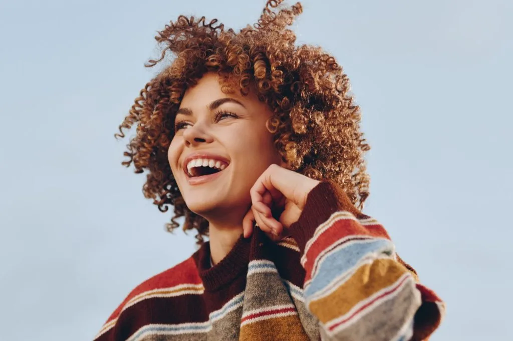 Woman with curly red hair smiling against blue sky wearing colorful striped sweater