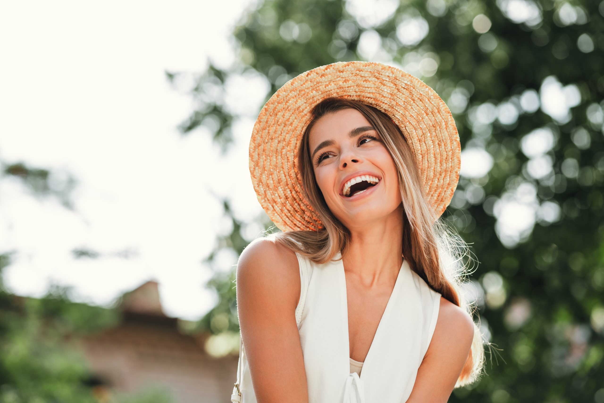 Woman wearing straw hat laughing outdoors under tree branches