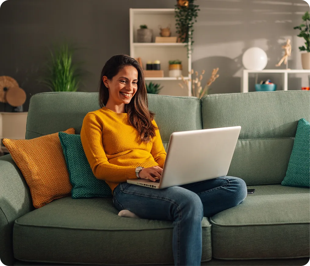 Woman in yellow sweater smiling while using a laptop on a green couch in a modern living room