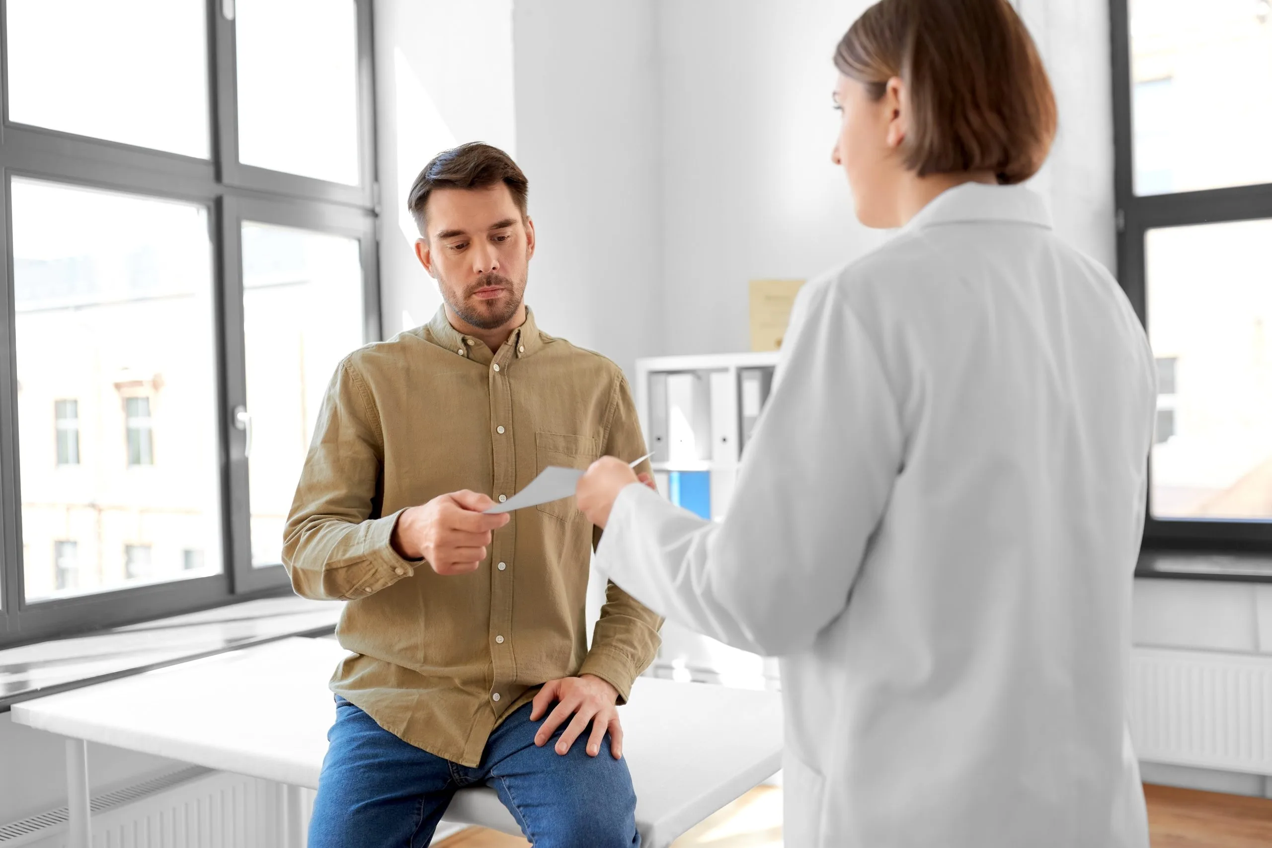 Doctor in white coat handing a prescription to a male patient in a bright clinical office