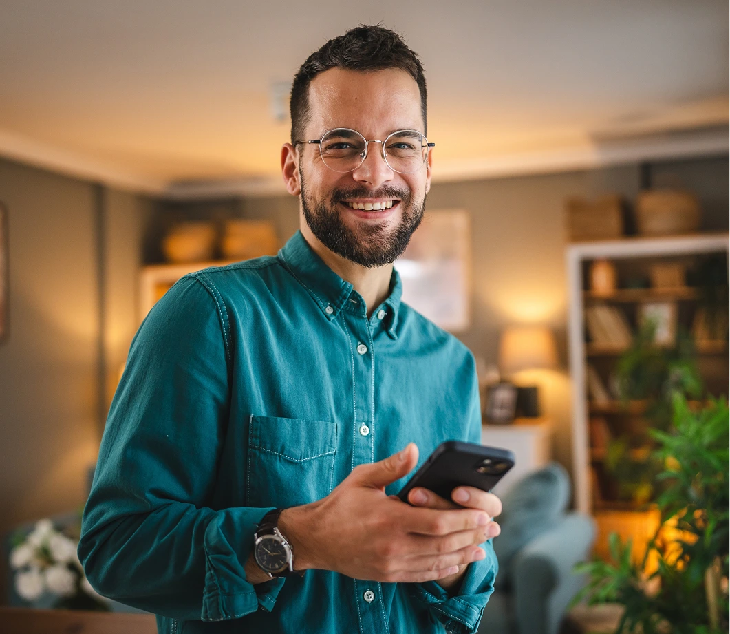 Man in teal shirt holding smartphone, smiling at camera in bright home office