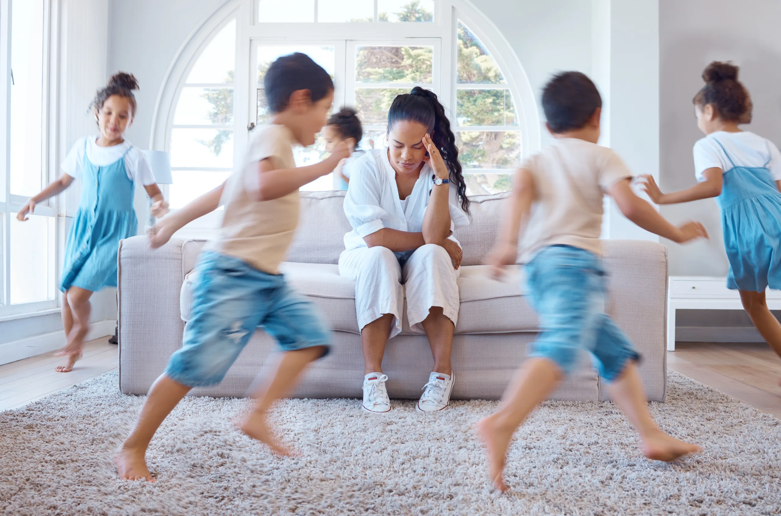Overwhelmed adult sitting on couch with hand to head while multiple children run around energetically in a bright living room
