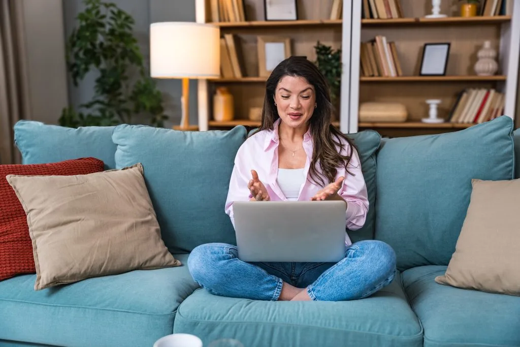 Woman sitting on a teal couch with a laptop, engaged in a telehealth session in a bright home office setting